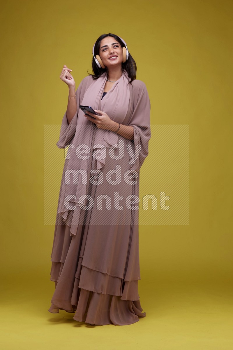 A Saudi woman Listing to Music on a Yellow Background wearing Brown Abaya