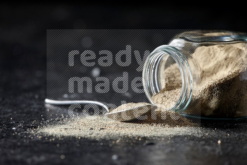 A flipped herbal glass jar and a metal spoon full of white pepper powder with spilled powder on textured black flooring