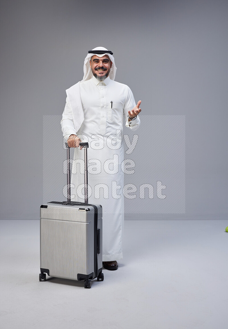 Saudi man wearing Thob and white Shomag standing holding Travel bag on Gray background