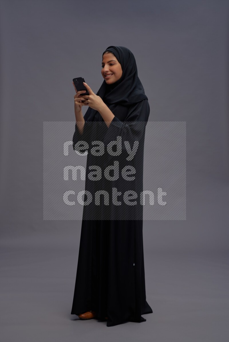 Saudi woman wearing Abaya standing texting on phone on gray background