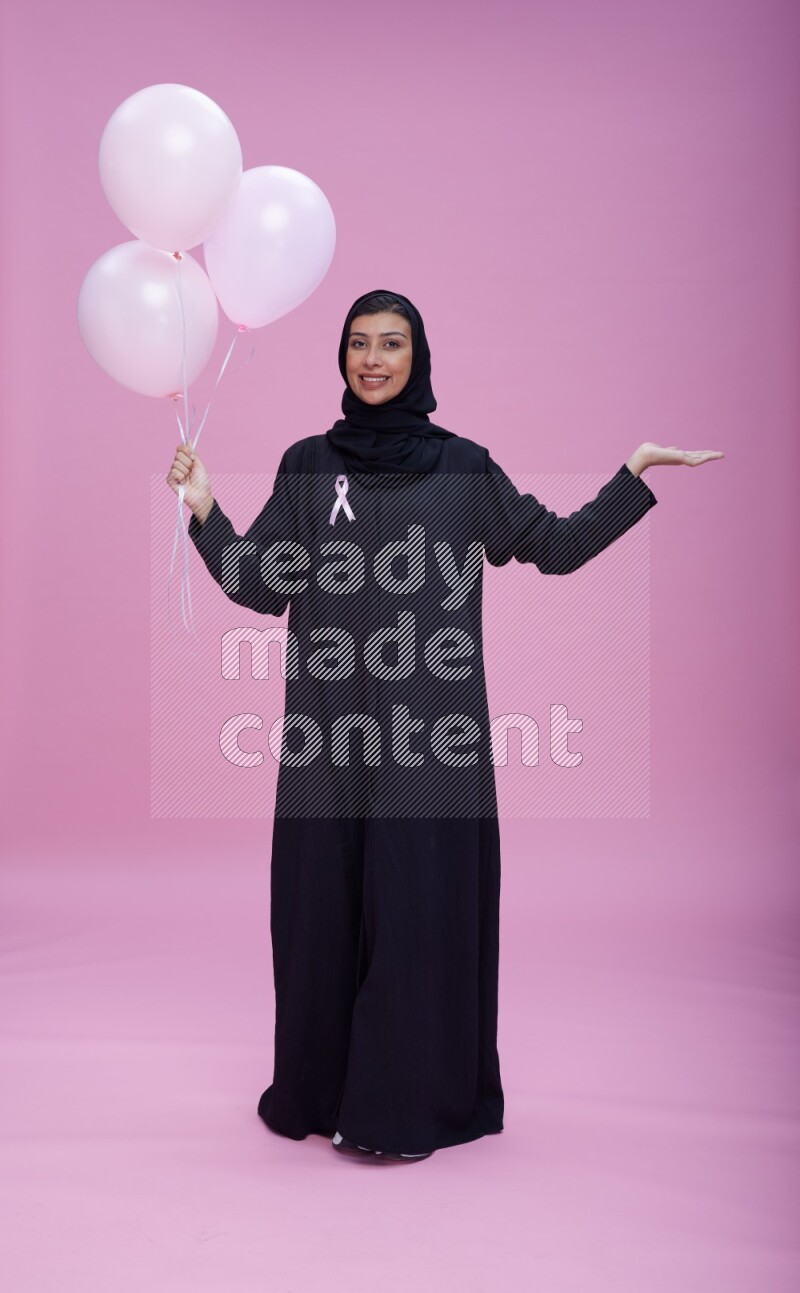 Saudi woman wearing pink ribbon on Abaya standing holding balloons on plain pink background