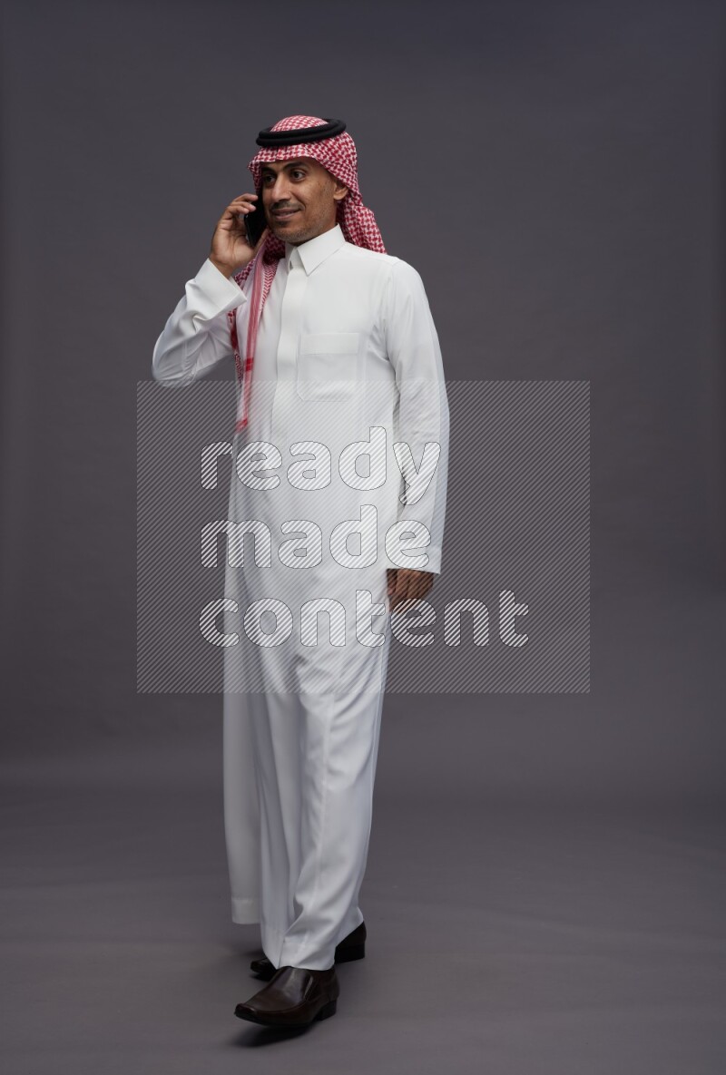 Saudi man wearing thob and shomag standing talking on phone on gray background