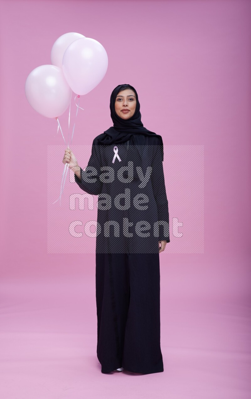 Saudi woman wearing pink ribbon on Abaya standing holding balloons on plain pink background