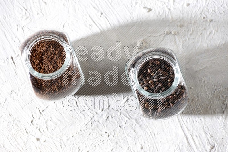 2 glass jars full of cloves powder and cloves grains on a textured white flooring