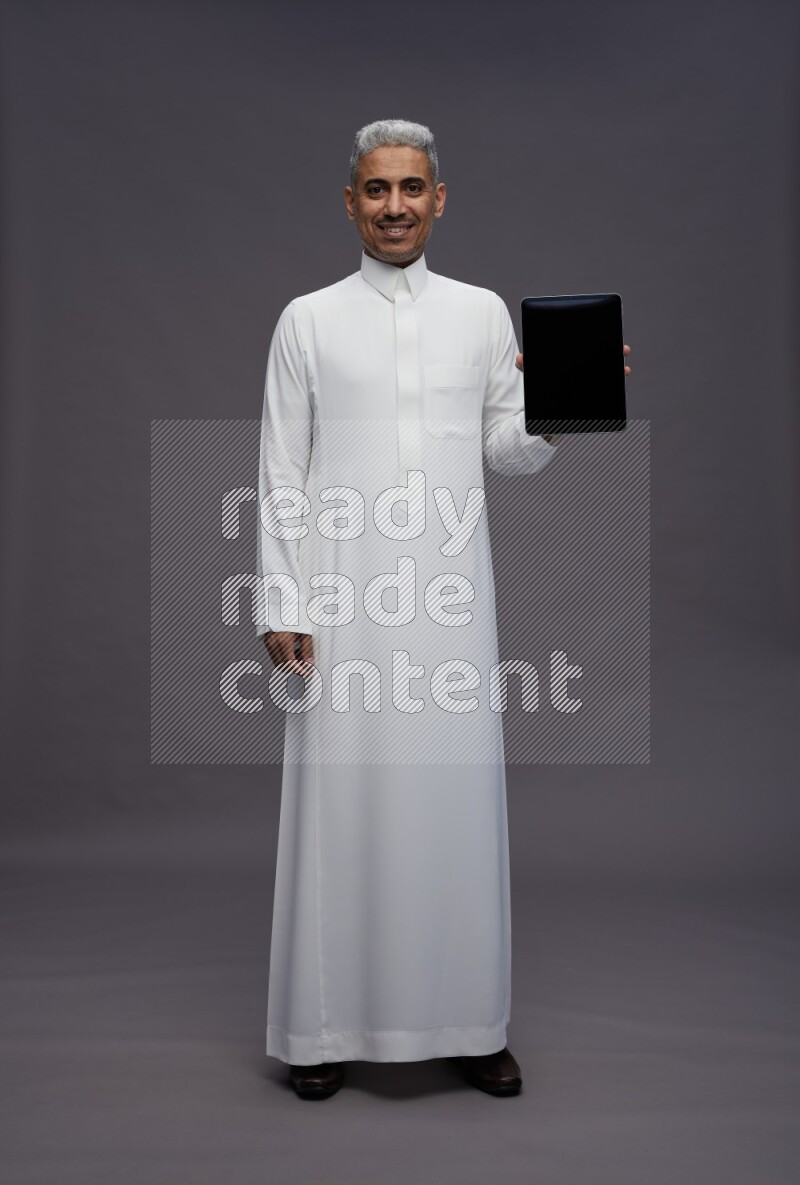 Saudi man wearing thob standing showing tablet to camera on gray background