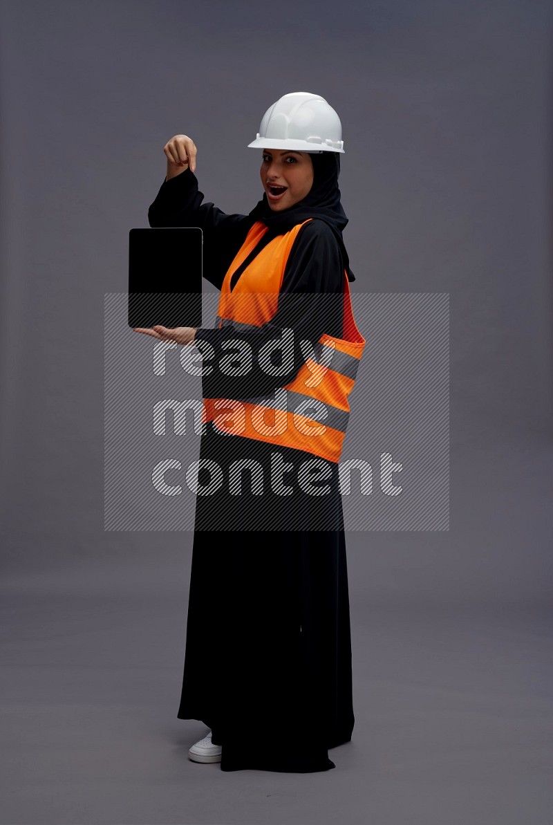 Saudi woman wearing Abaya with engineer vest standing showing tablet to camera on gray background