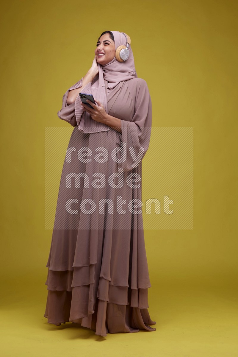 A woman Listening to Music on a Yellow Background wearing Brown Abaya with Hijab