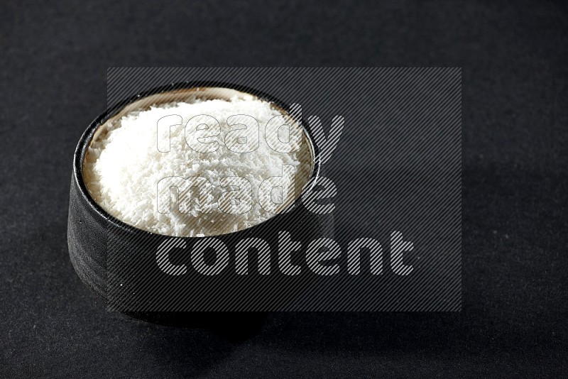 A black ceramic bowl full of desiccated coconut on a black background in different angles