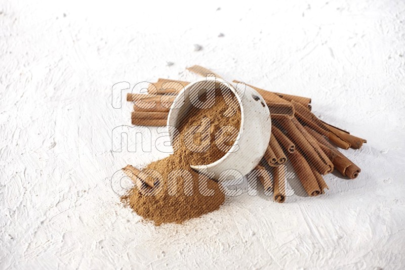 Ceramic beige bowl over filled with cinnamon powder and cinnamon sticks around the bowl on a textured white background in different angles