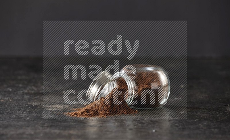 A flipped glass spice jar full of cloves powder and powder came out of it on textured black flooring