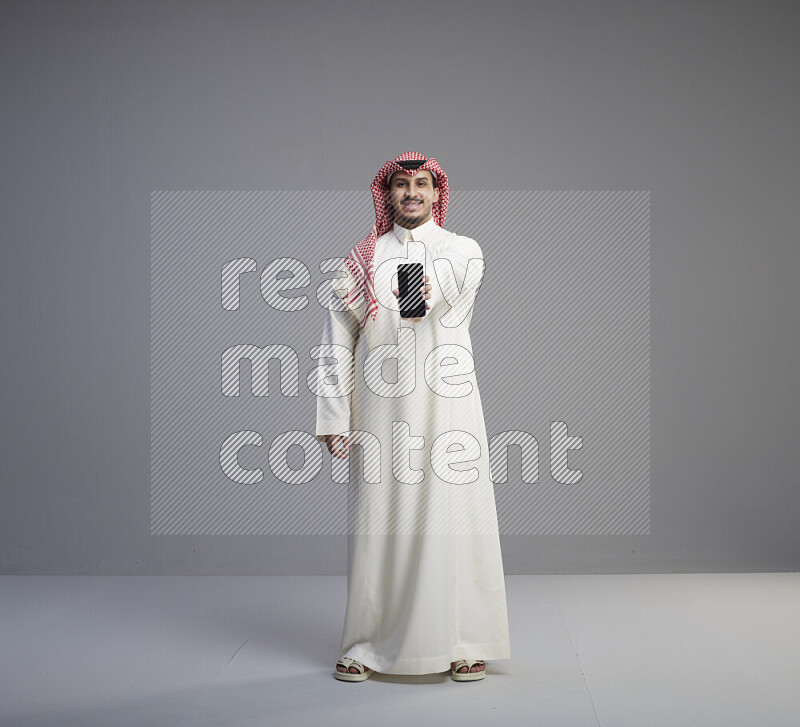 A Saudi man standing wearing thob and red shomag showing phone to camera on gray background