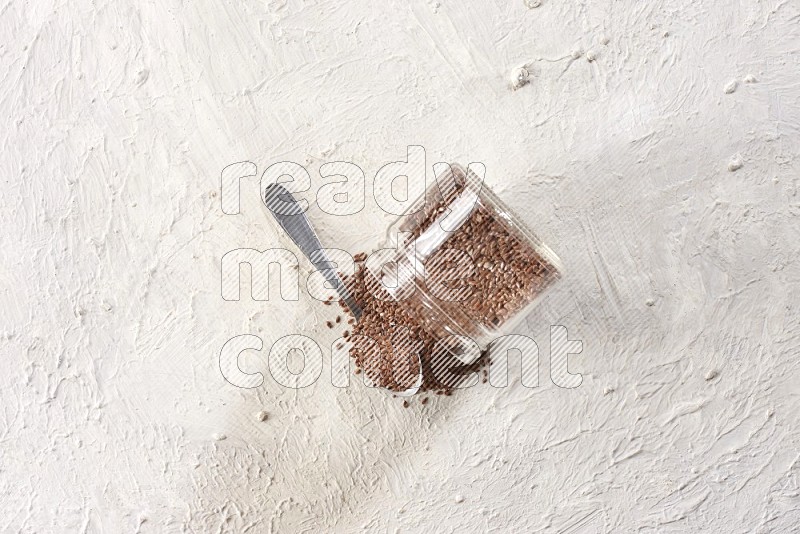 A glass jar full of flax seeds flipped and seeds spread out with a metal spoon full of the seeds on a textured white flooring