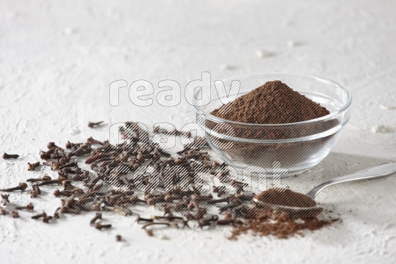 A glass bowl and a metal spoon full of cloves powder with cloves grains spread on a textured white flooring