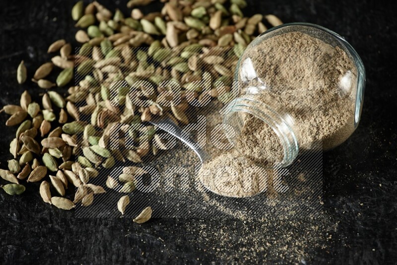 وعاء زجاجي للتوابل مقلوب وملعقة معدنية ممتلئة ببودرة الهيل وحبوب الهيل متناثرة على خلفية سوداء