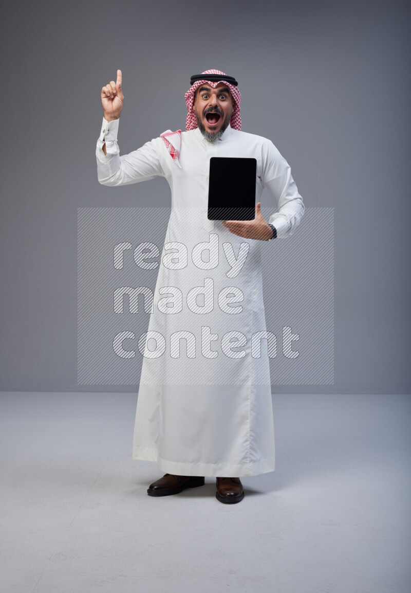 Saudi man Wearing Thob and red Shomag standing showing tablet to camera on Gray background
