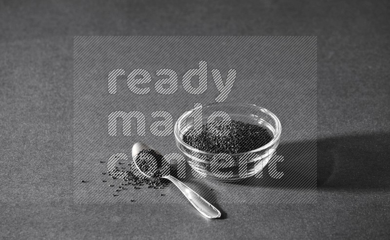 A glass bowl full of black seeds with a metal spoon full of the seeds on a black flooring