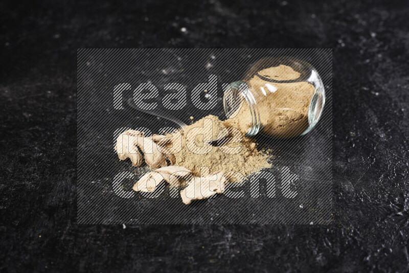 A glass jar full of ground ginger powder flipped with some spilling powder on black background