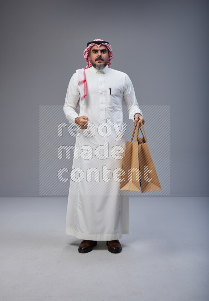 Saudi man Wearing Thob and red Shomag standing holding shopping bag on Gray background