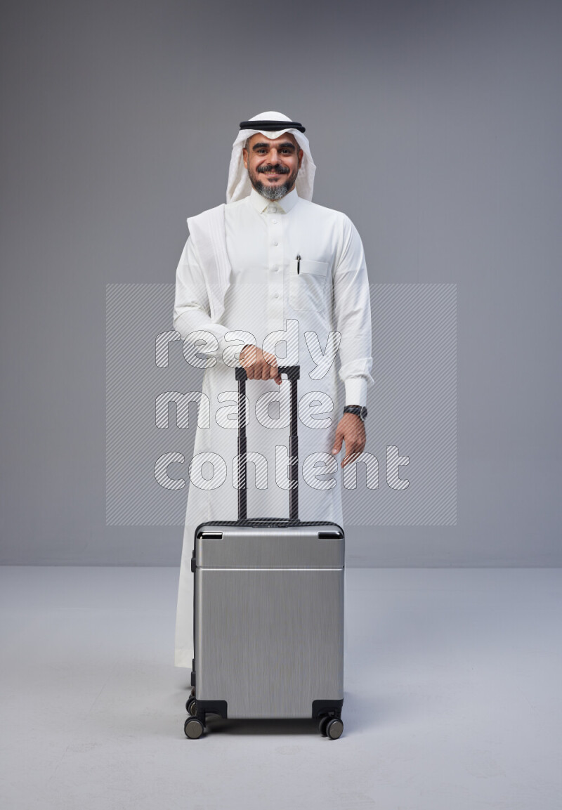 Saudi man wearing Thob and white Shomag standing holding Travel bag on Gray background