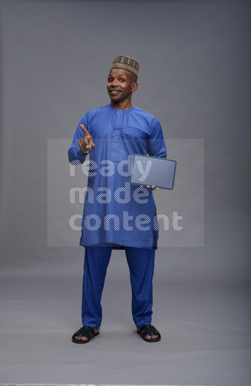 Man wearing Nigerian outfit standing showing tablet to camera on gray background