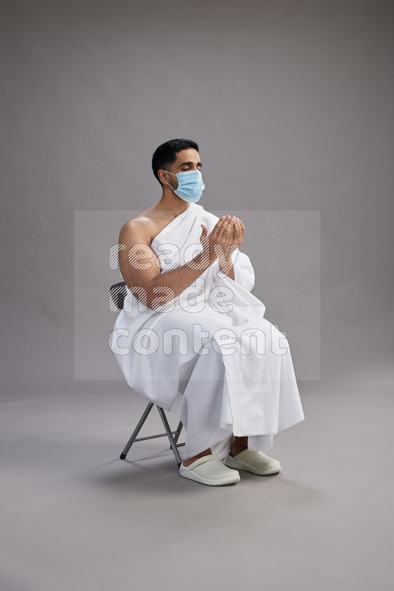 A man wearing Ehram with face mask sitting on chair dua'a on gray background
