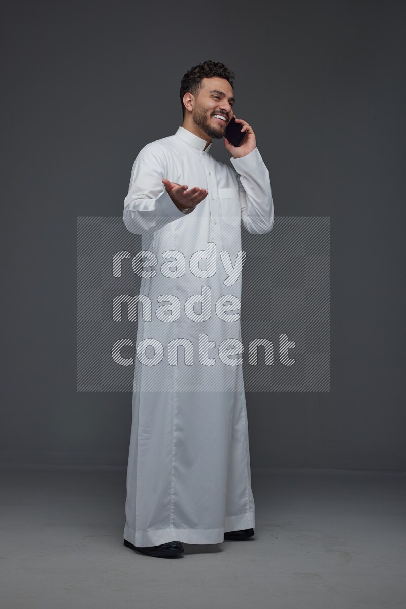 A Saudi man wearing Thobe and talking in the phone while standing and making different poses eye level on a gray background
