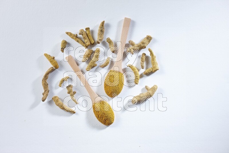 2 wooden spoons full of turmeric powder with dried turmeric fingers beside it on white flooring