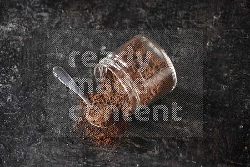 A flipped glass jar and metal spoon full of cloves powder with cloves spread on a textured black flooring