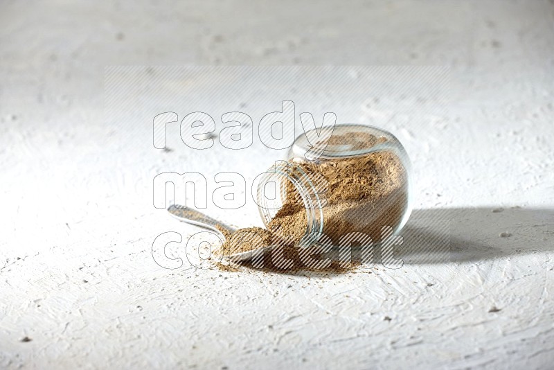 A flipped glass spice jar and a metal spoon full of cumin powder and powder spilled out on textured white flooring