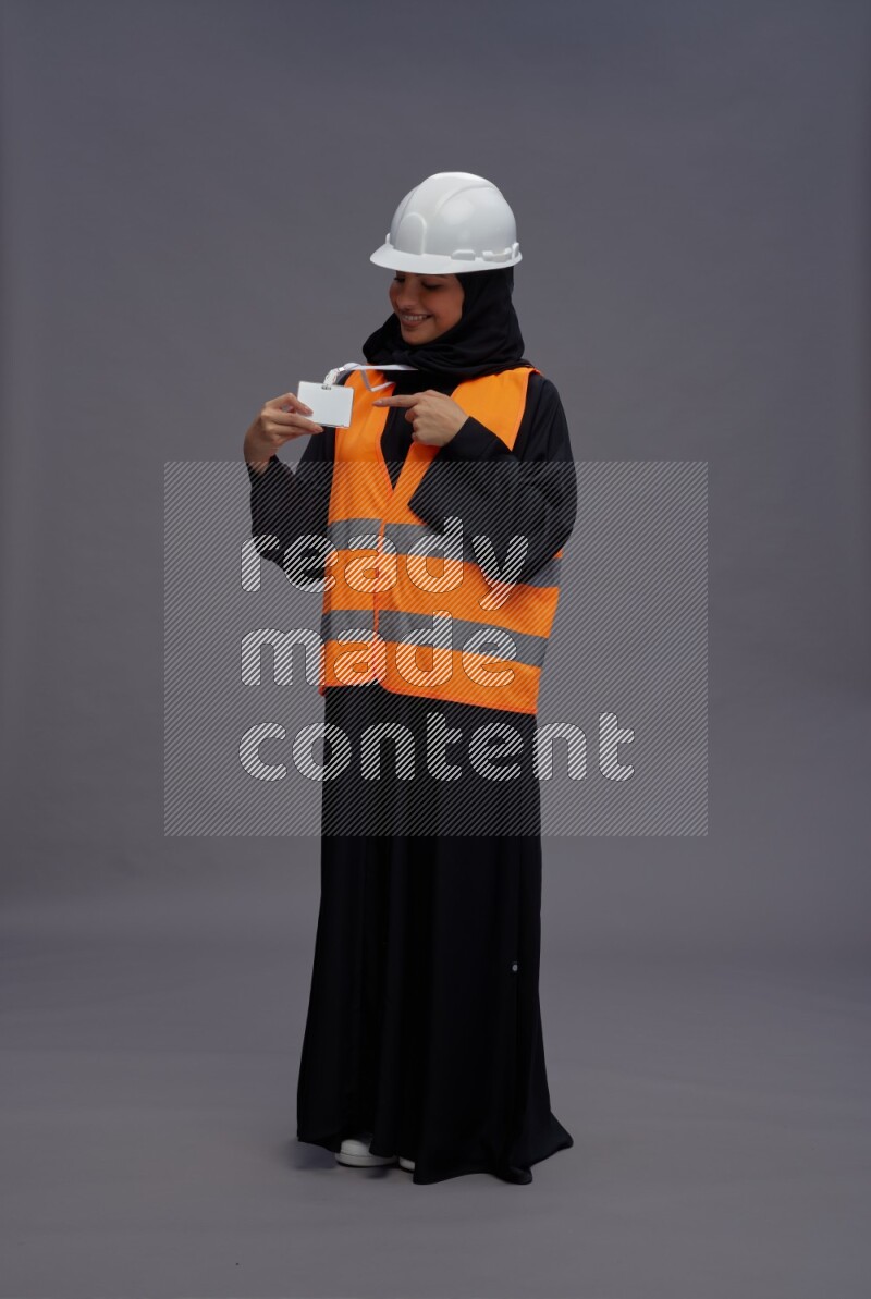 Saudi woman wearing Abaya with engineer vest with neck strap employee badge standing interacting with the camera on gray background