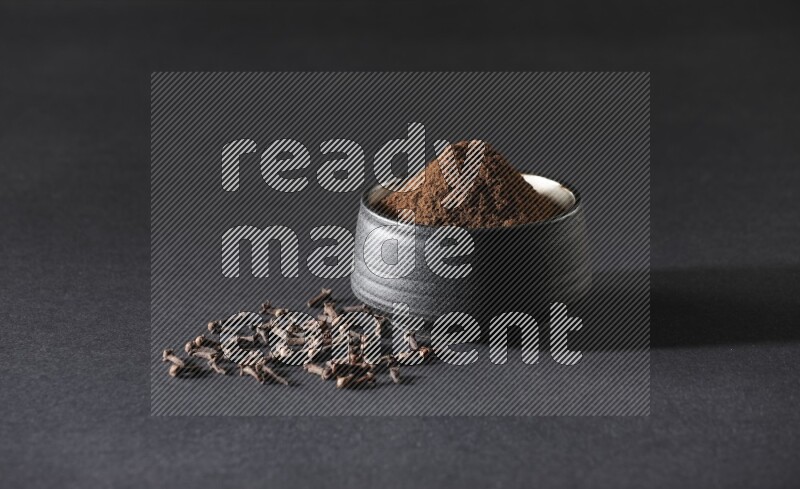 A black pottery bowl full of cloves powder and cloves whole grains on a black flooring