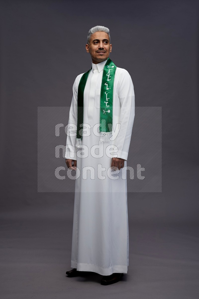 Saudi man wearing thob standing holding Saudi flag on gray background