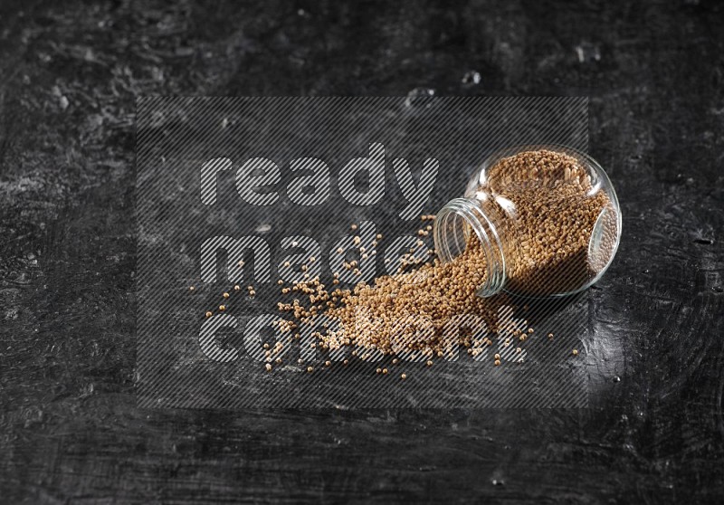 A glass spice jar full of mustard seeds and jar is flipped and seeds spread out on a textured black flooring