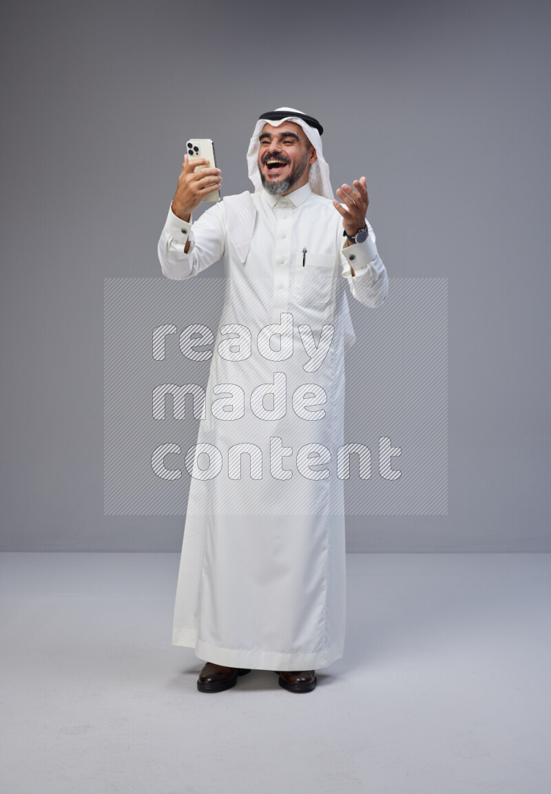 Saudi man Wearing Thob and white Shomag standing texting on phone on Gray background