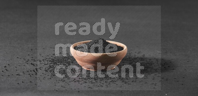 A wooden bowl full of black seeds surrounded by seeds on a black flooring