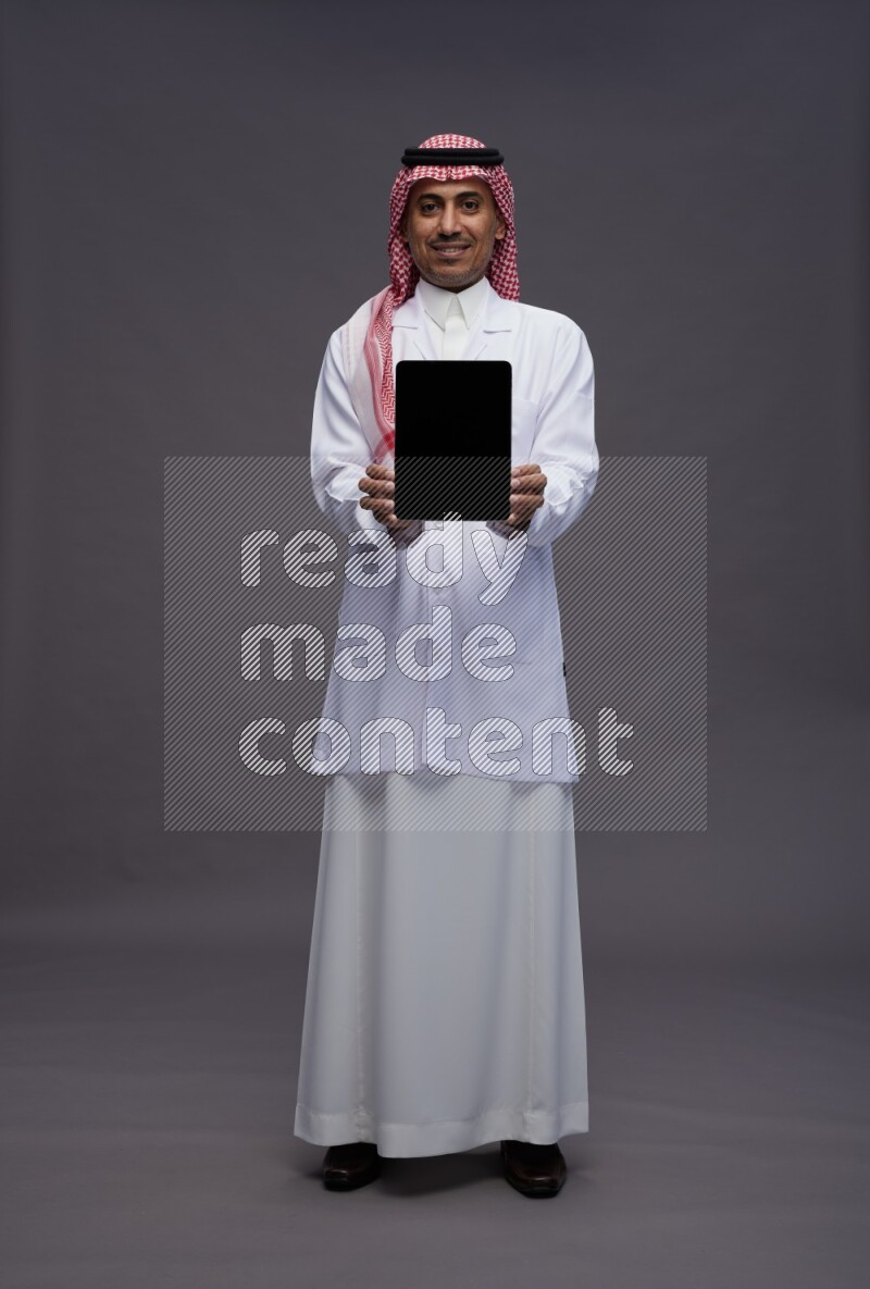 Saudi man wearing thob with lab coat and shomag with pocket employee badge standing showing tablet to camera on gray background