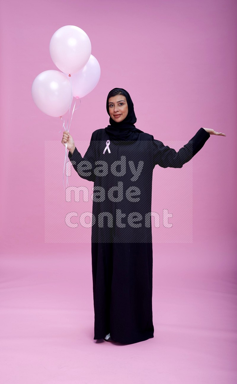 Saudi woman wearing pink ribbon on Abaya standing holding balloon on plain pink background