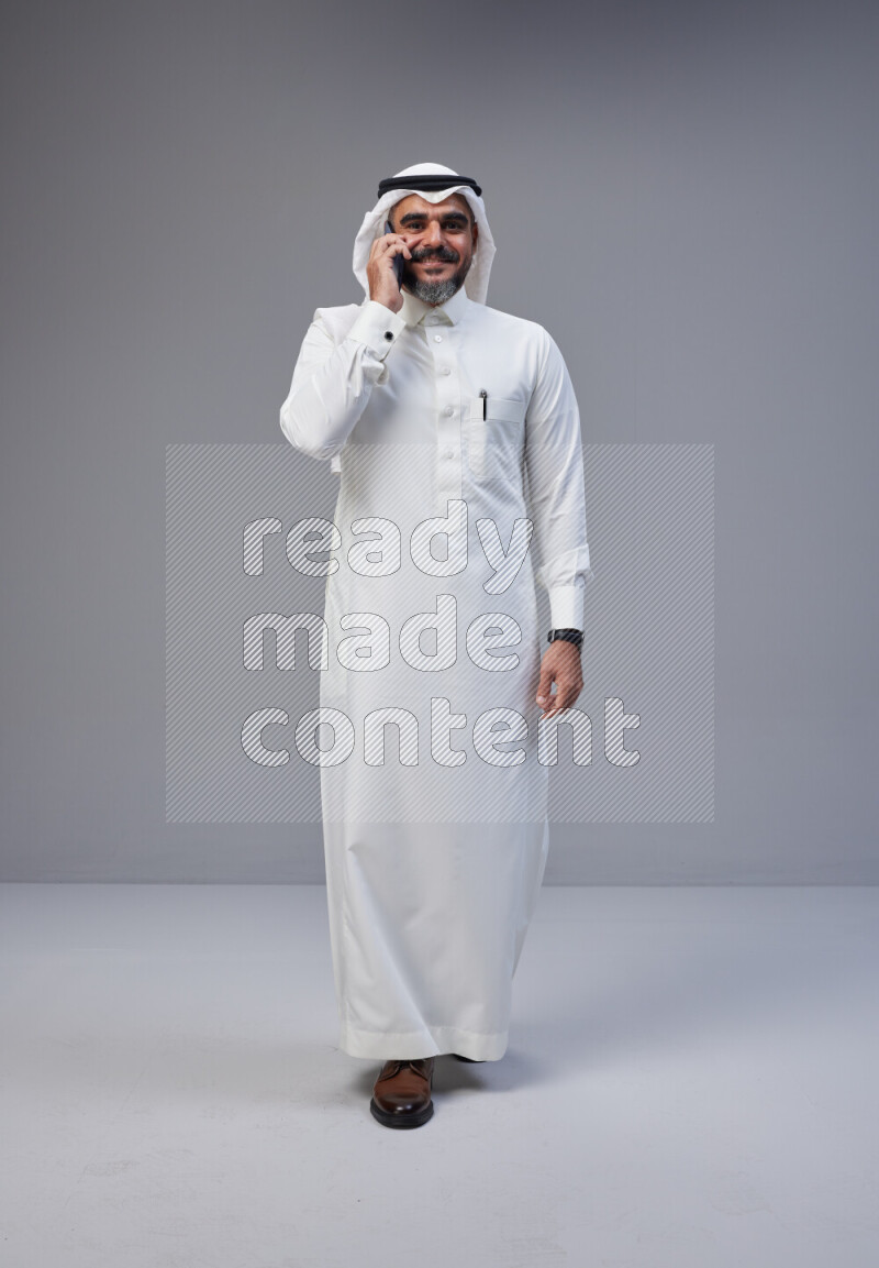 Saudi man Wearing Thob and white Shomag standing talking on phone on Gray background