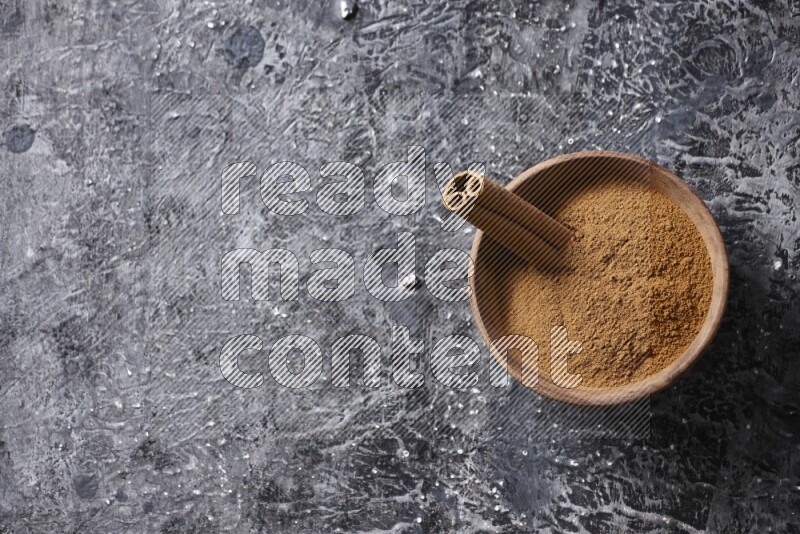 Wooden bowl full of cinnamon powder and a cinnamon stick on a textured black background