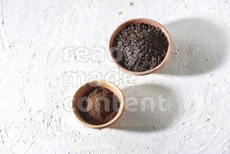 2 wooden bowls full of cloves powder and whole cloves on a textured white flooring