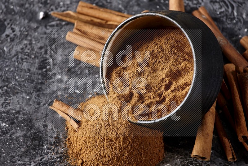 Black pottery bowl over filled with cinnamon powder and cinnamon sticks around the bowl on a textured black background