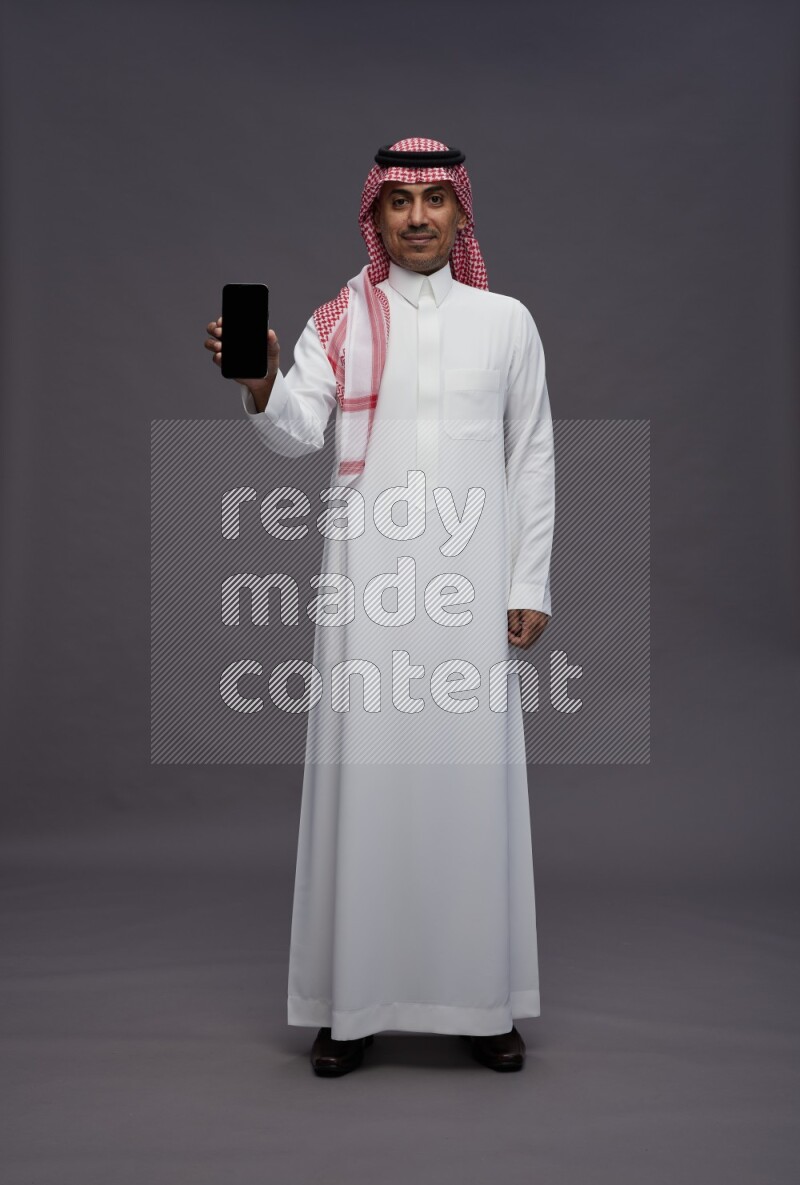 Saudi man wearing thob and shomag standing showing phone to camera on gray background