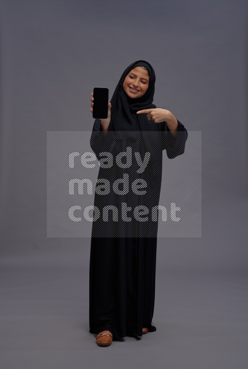 Saudi woman wearing Abaya standing showing phone to camera on gray background