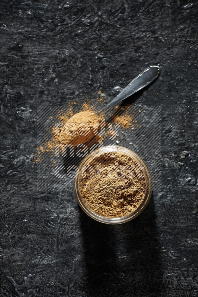 A glass jar and a metal spoon full of allspice powder on a textured black flooring