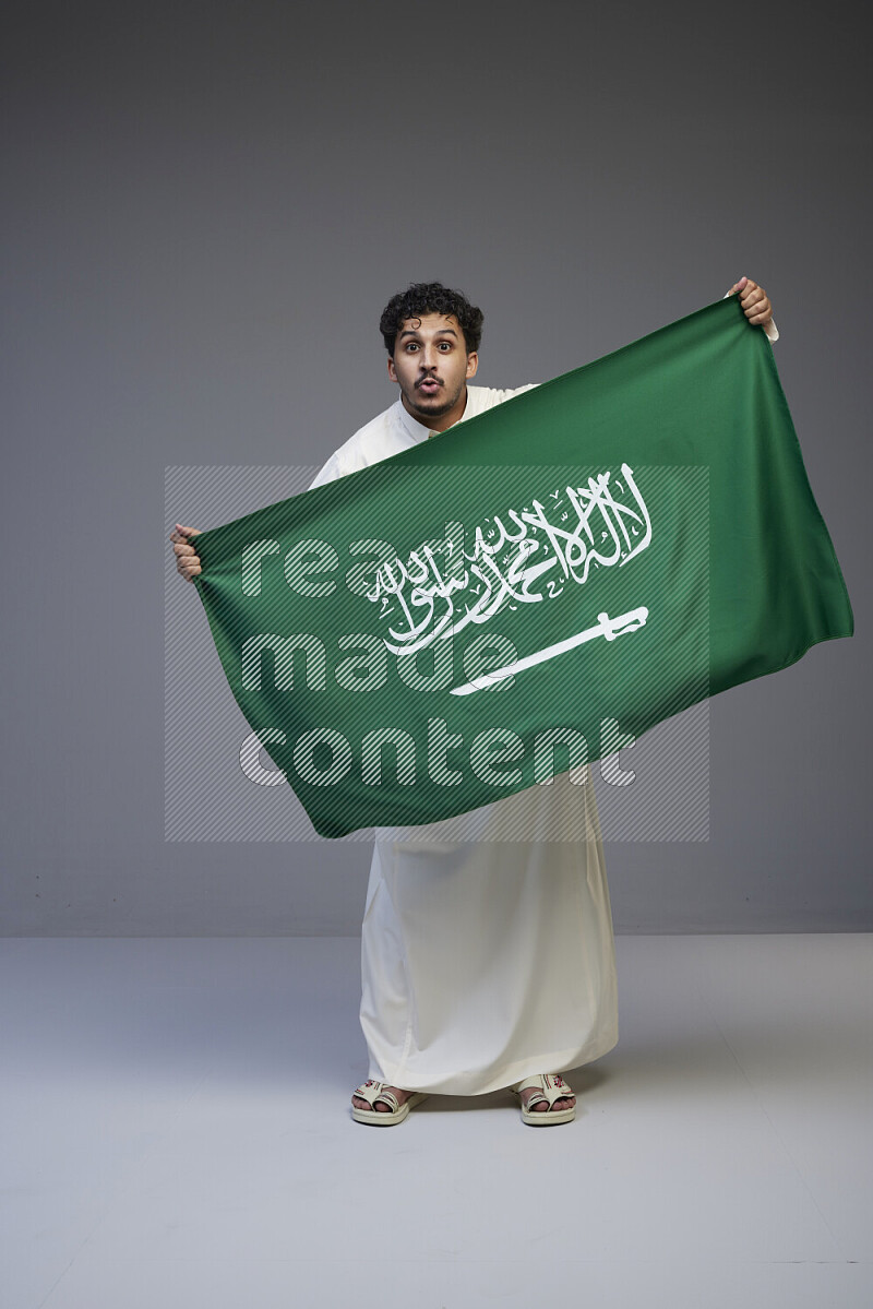 A Saudi man standing wearing thob holding big Saudi flag on gray background