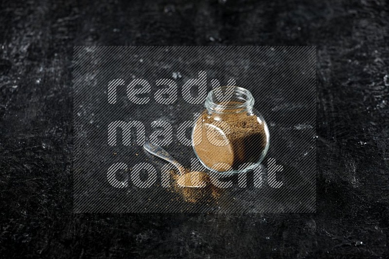 A glass spice jar and a metal spoon full of cumin powder on a textured black flooring