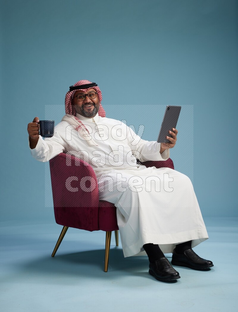 Saudi Man with shimag sitting on chair working on tablet on blue background