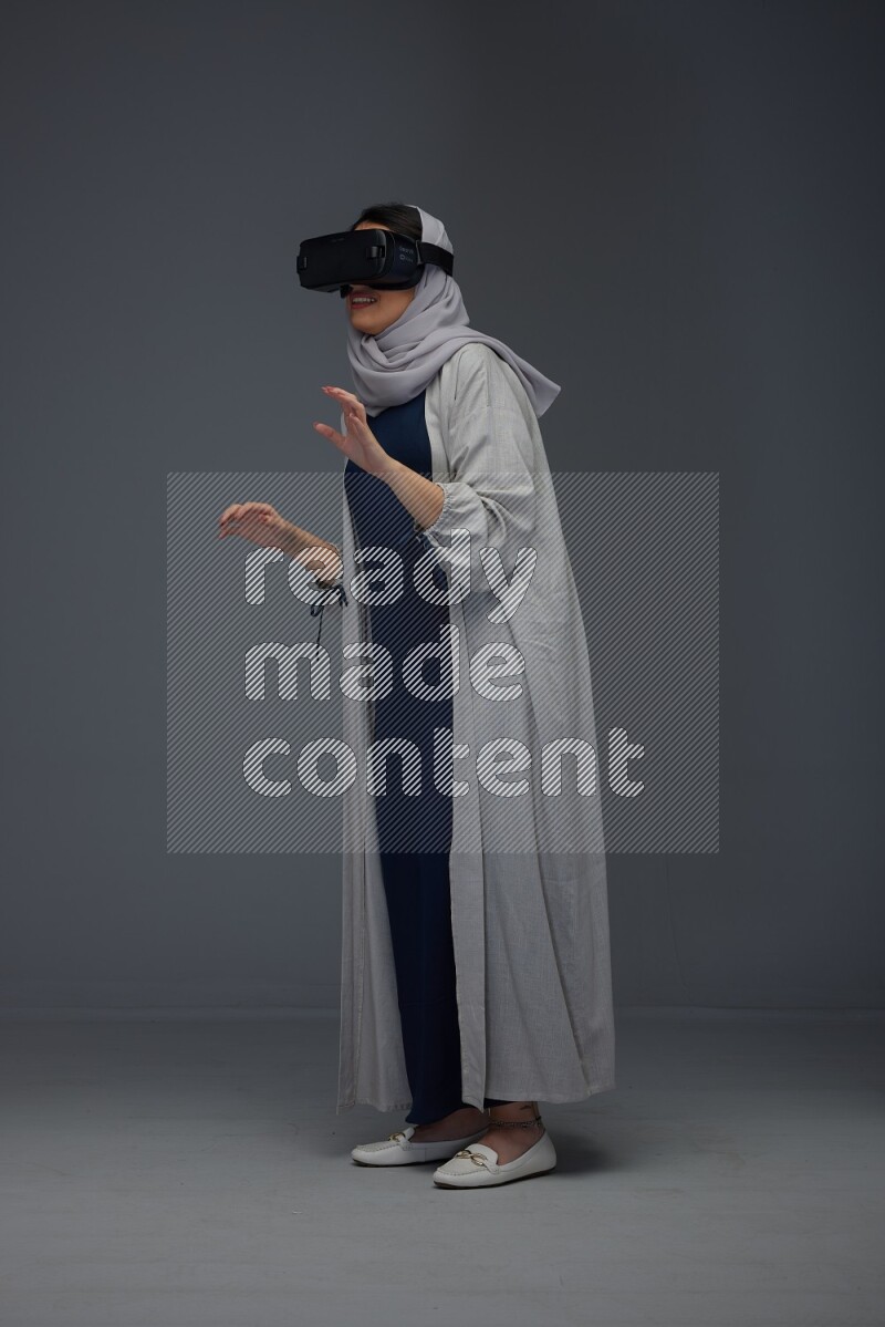 A Saudi woman wearing a light gray Abaya and head scarf standing and wearing VR eye level on a grey background