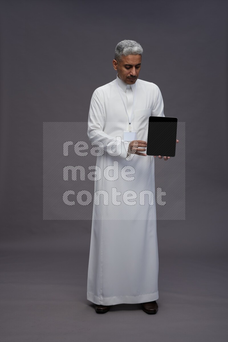 Saudi man wearing thob with neck strap employee badge standing showing tablet to camera on gray background