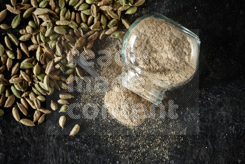 وعاء زجاجي للتوابل مقلوب وملعقة معدنية ممتلئة ببودرة الهيل وحبوب الهيل متناثرة على خلفية سوداء